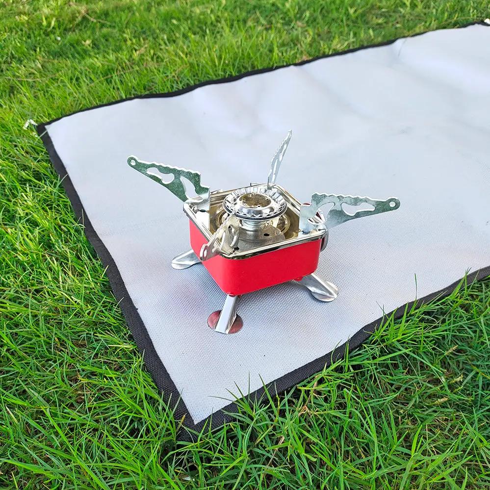 Camping fireproof cloth grill mat, gray with black edge, laid on grass beneath a small portable stove
