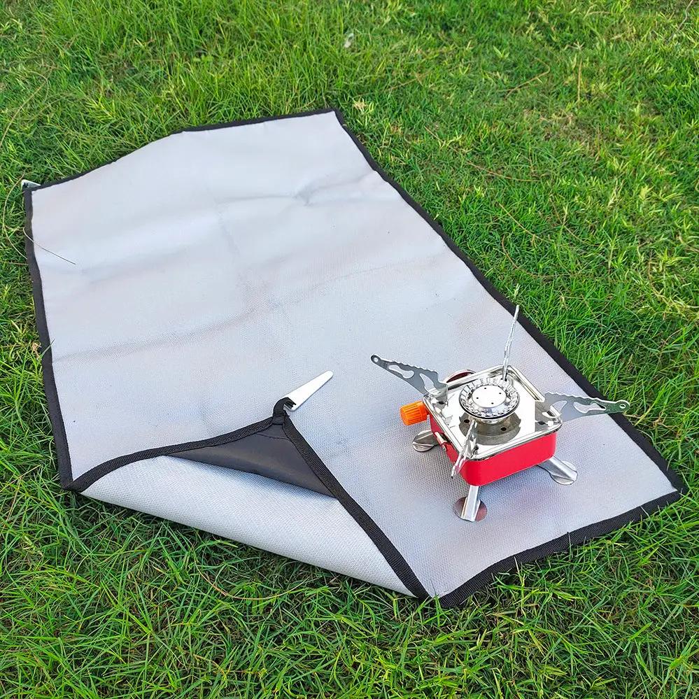 Camping grill mat, gray flame-retardant with black border on grass; 1 red portable stove beside it.