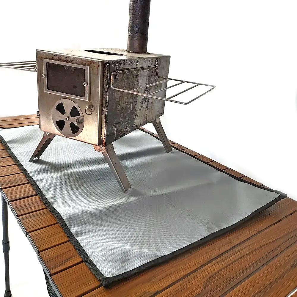 Fireproof BBQ grill mat, light gray flame-retardant cloth under a camping stove on a wooden deck.