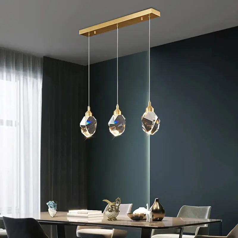 Three crystal pendant lamps with gold fittings hanging from a gold ceiling bar over a modern dining table