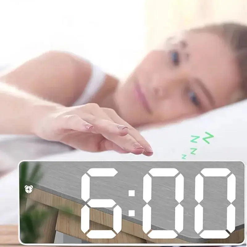 Acrylic mirror digital alarm clock on a bedside table displaying 6:00 in a dim bedroom.