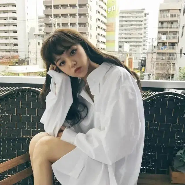 White oversized women's button-up shirt with long sleeves worn by a model on a balcony, minimalist chic style.