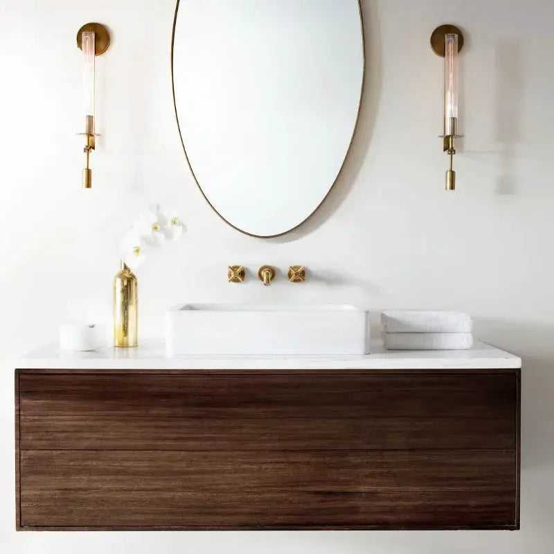 Modern copper wall sconces with glass tubes flank an oval mirror above a white sink on a wooden vanity