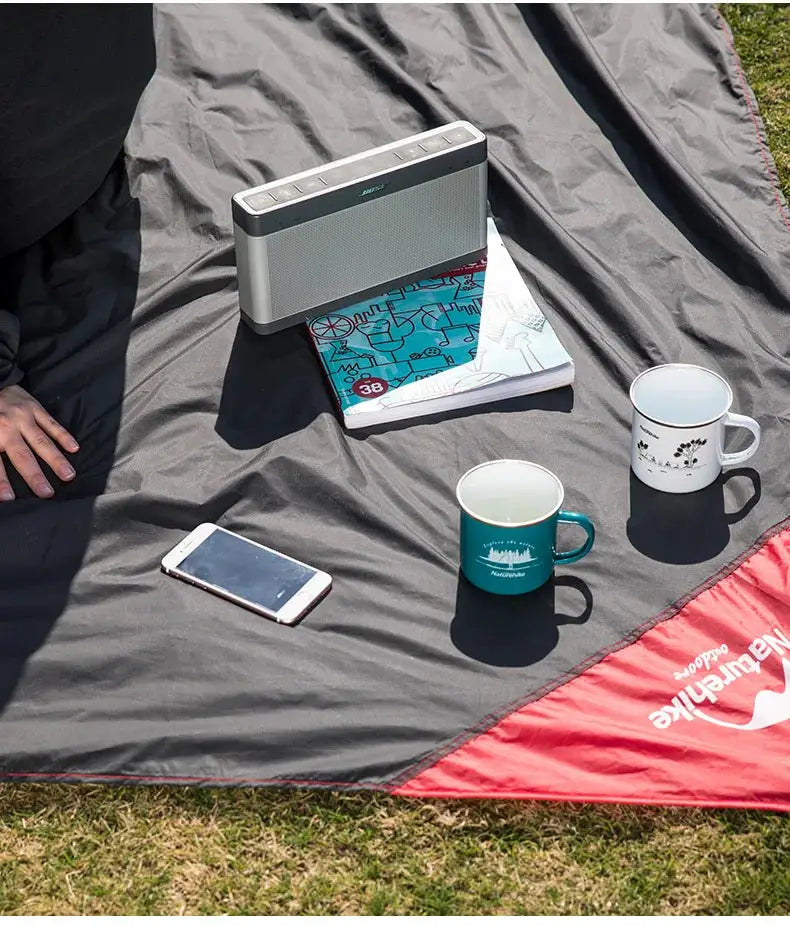 Naturehike picnic blanket gray waterproof mat with red edge on grass; includes portable speaker, two mugs, and phone
