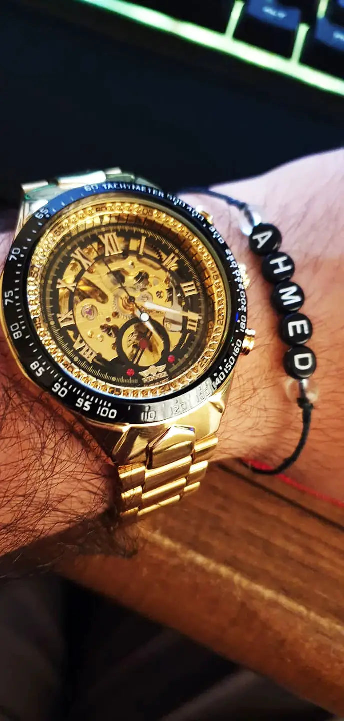 Gold-tone men's mechanical skeleton watch with black bezel, Roman numeral markers, and metal bracelet