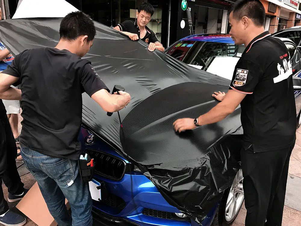 Car vinyl wrap sheet 4D carbon fiber applied to a blue BMW hood and roof by three workers.