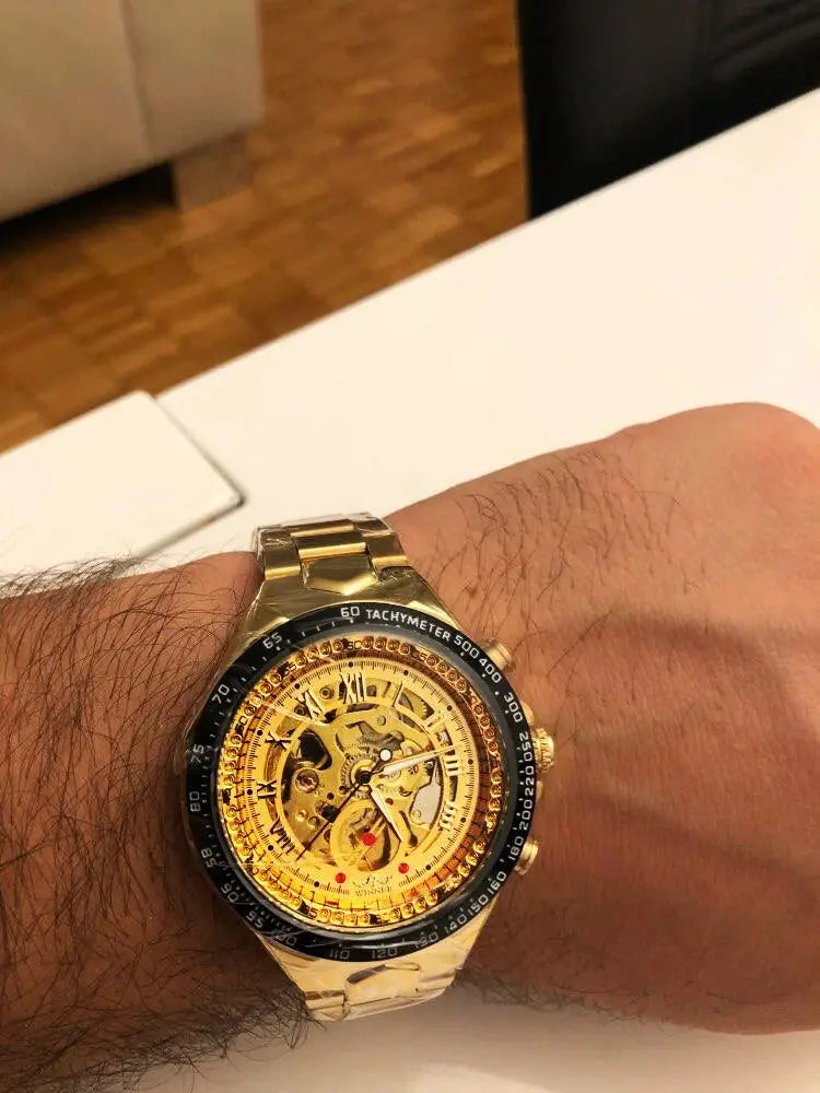 Men's skeleton mechanical sport watch on wrist with black tachymeter bezel and gold bracelet.