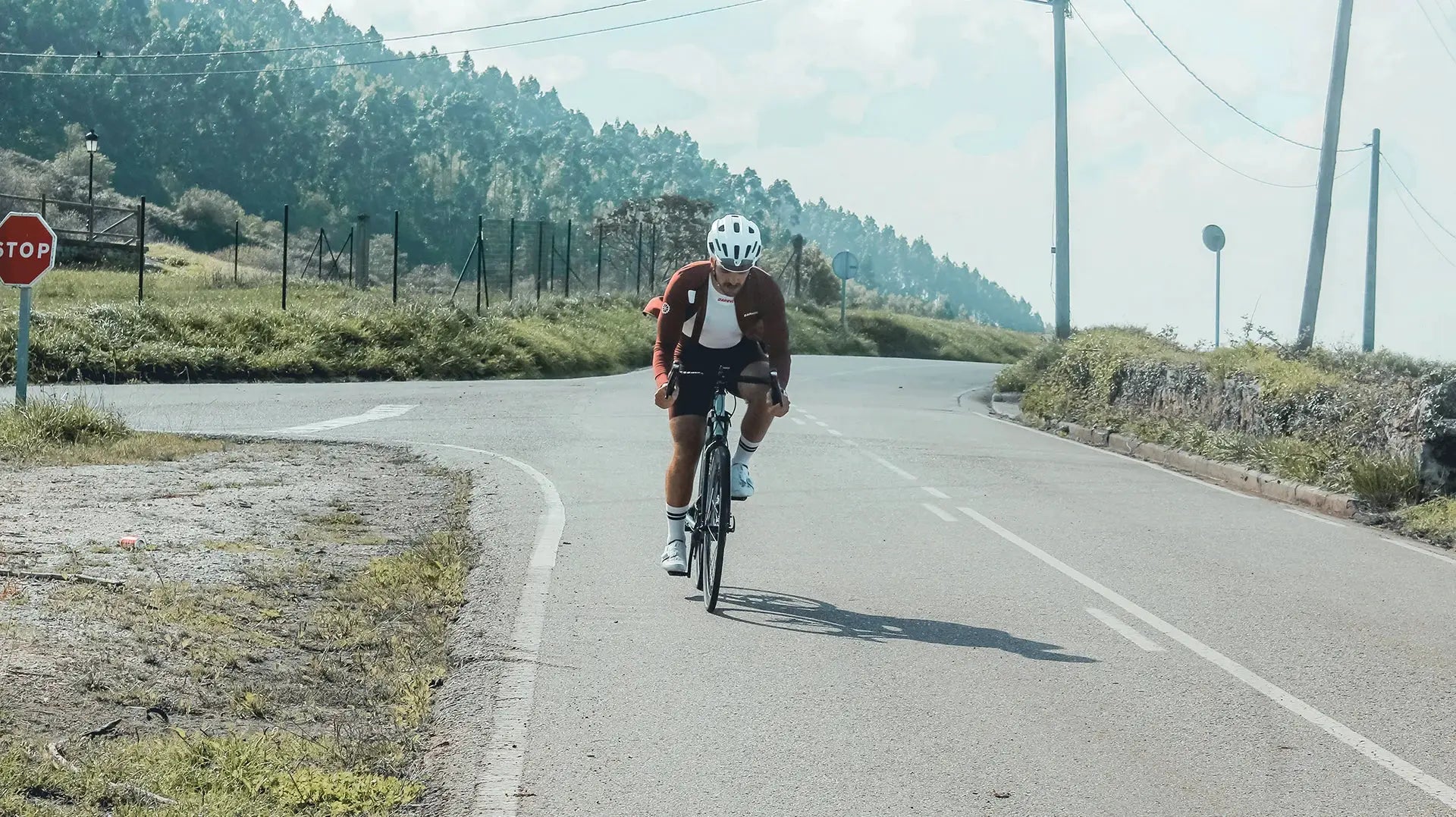 DAREVIE cycling jersey on male rider in maroon, white helmet, riding a road bike on a winding rural road.