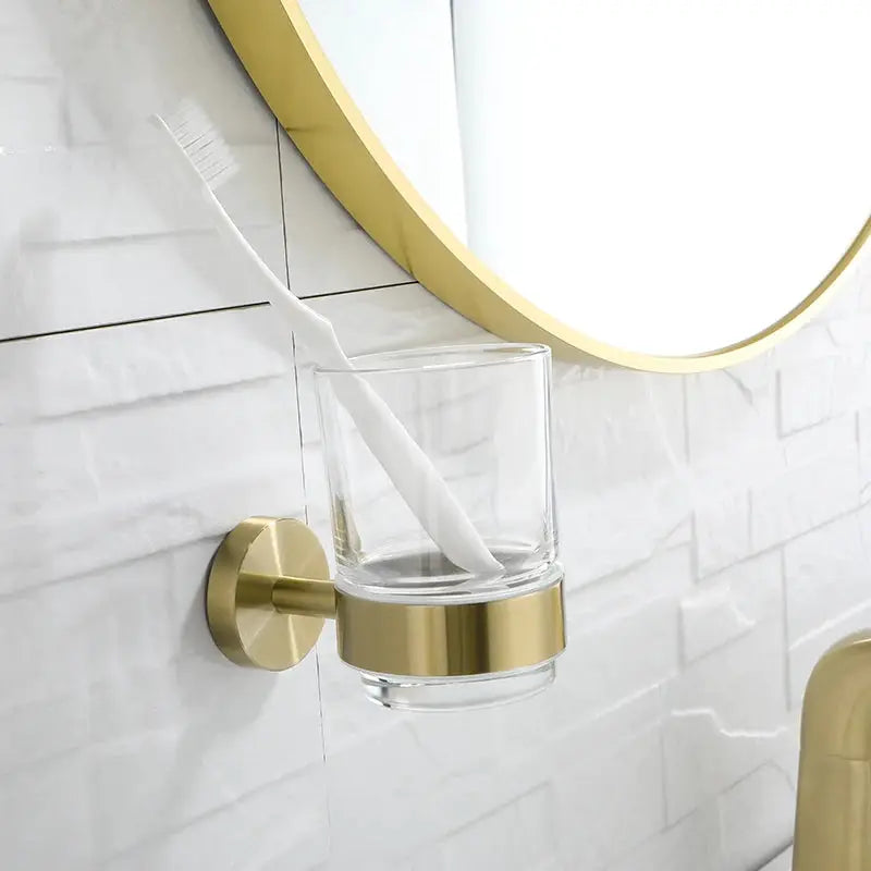 Toothbrush holder with glass tumbler in brushed gold, wall-mounted bathroom accessory.