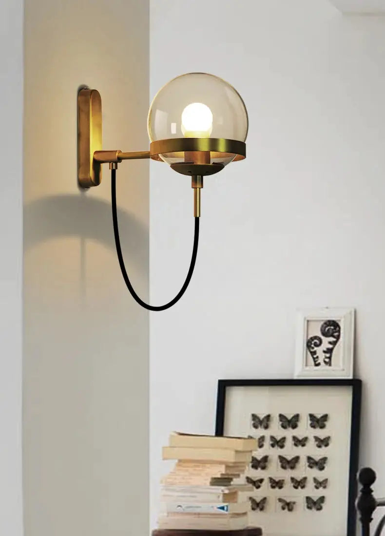 Modern glass ball wall lamp by Simples Market; brass finish, clear globe, black cord.