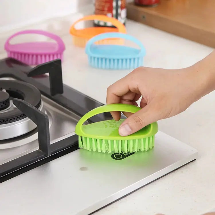 Green egg-shaped cleaning brush with soft bristles used on a stovetop; colorful brushes in the background.