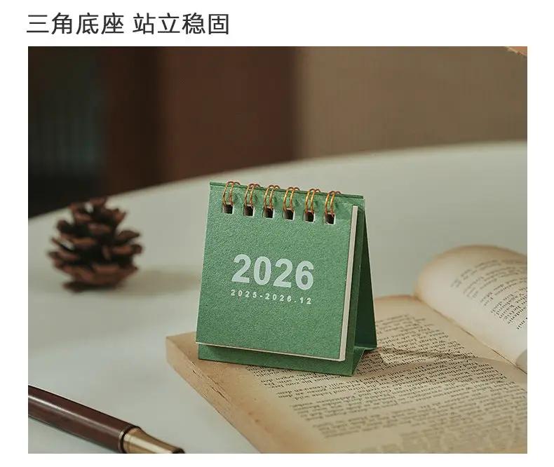 Mini green 2026 desk calendar with coil binding from Simples Market, on a desk beside an open book