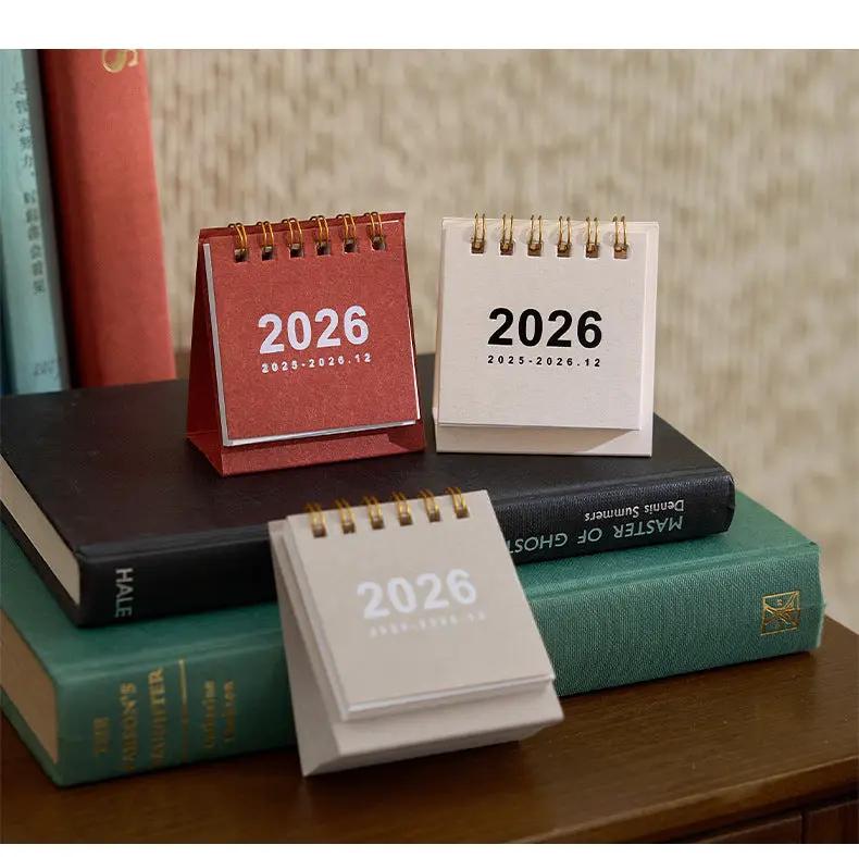 Simples Market 2026 mini desk calendar set; three coil-bound calendars in red, beige and gray atop stacked books.