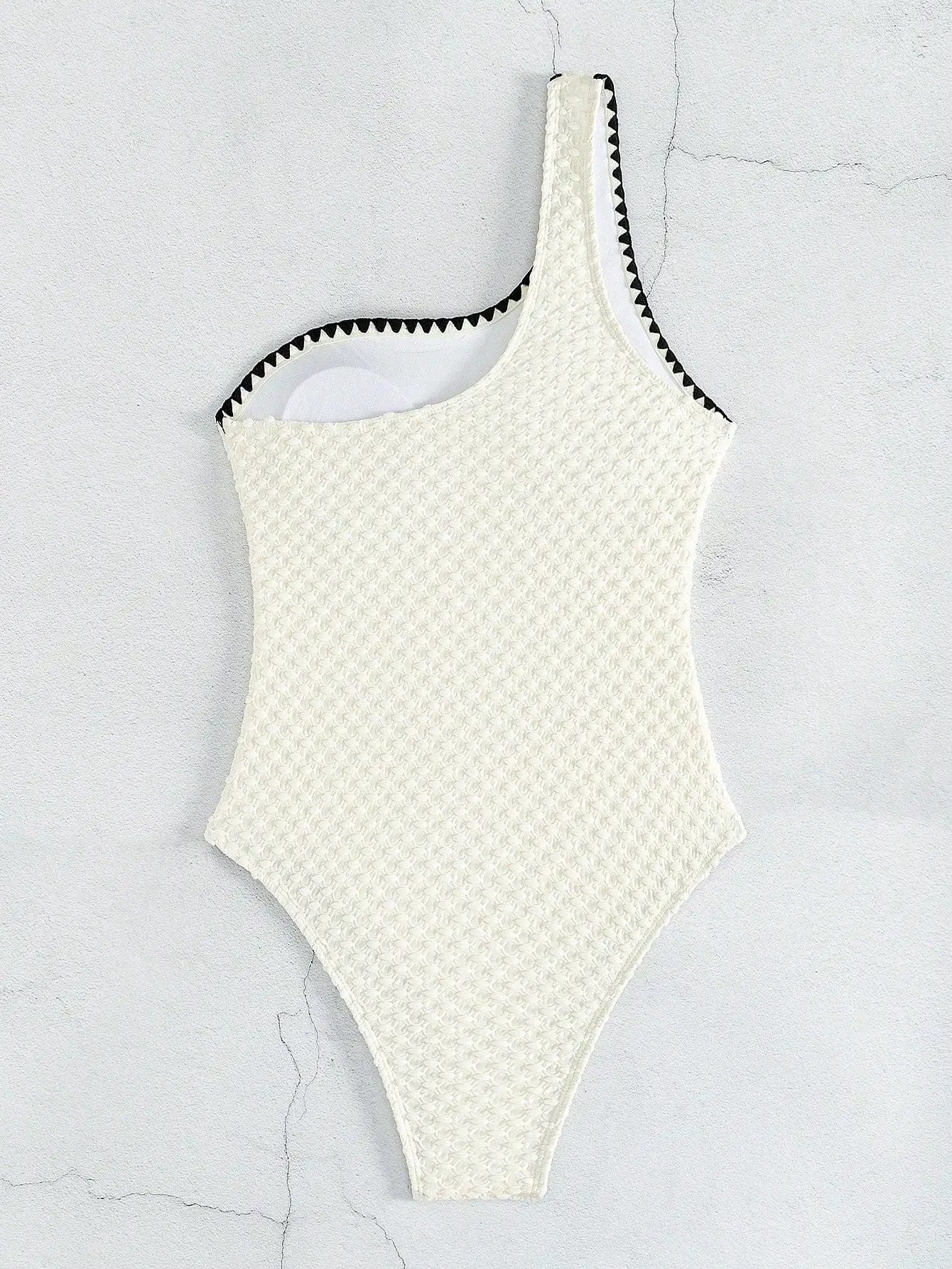 Simples Market white textured one-piece swimsuit with a one-shoulder strap and black zigzag trim.