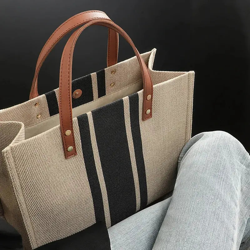 Canvas tote bag with beige body, central black striped panel, white accents, large capacity, brown leather top handles