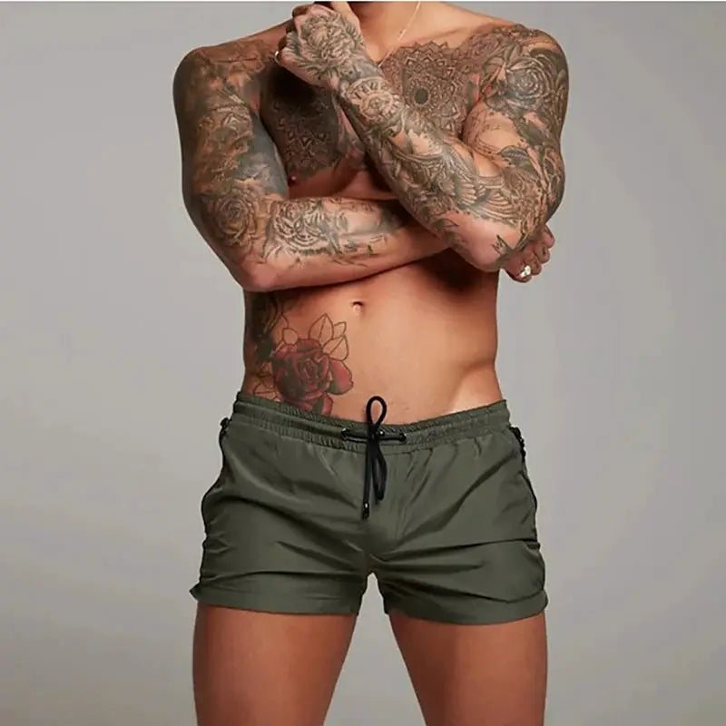 Men's olive green swim trunks with black drawstring on a tattooed model.