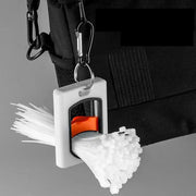 Square white cable tie organizer with orange slider; stores white zip ties and attaches to a backpack via metal ring.