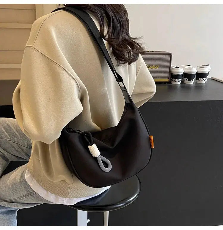 Crossbody bag in black with a wide shoulder strap, dumpling-style silhouette and white rope loop accent.
