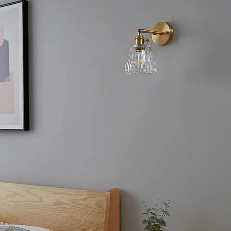 Brass glass wall lamp with a ribbed shade mounted on a gray wall above a wooden headboard.
