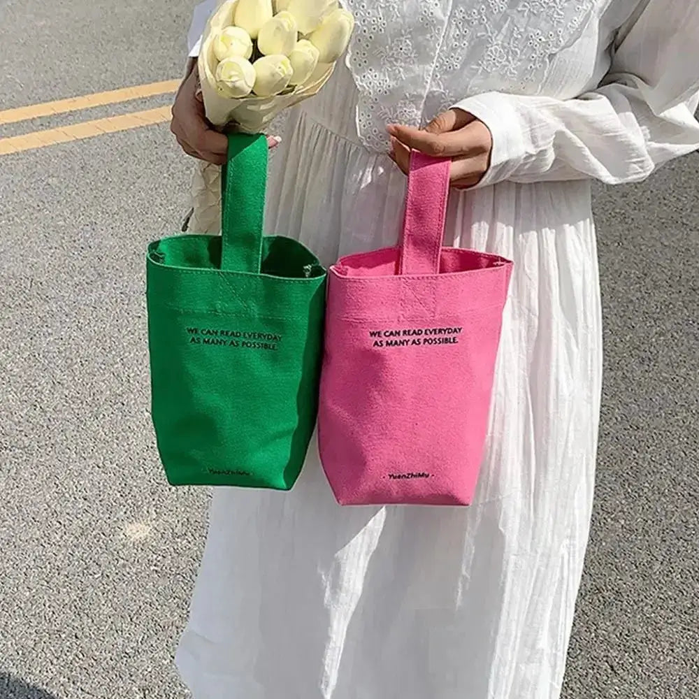 Two canvas crossbody bags in green and pink with short handles and printed slogan.