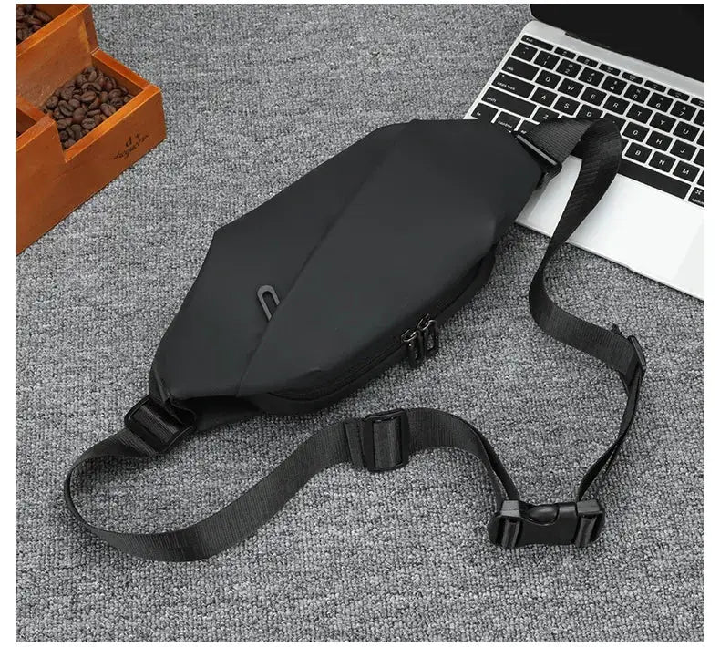 Black waist bag with adjustable strap and dual zippers, placed beside a laptop on a gray carpet
