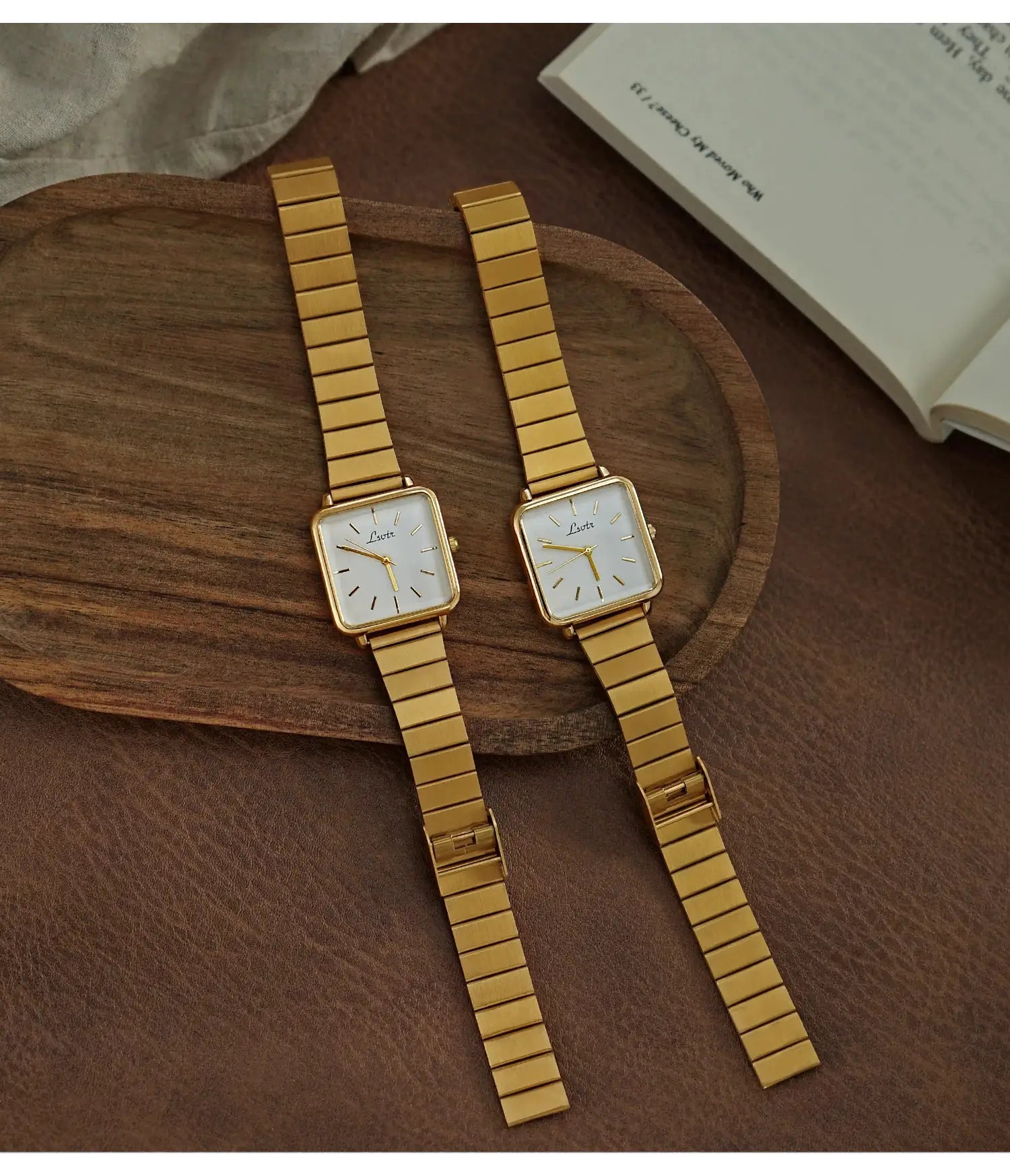 Two gold-tone square-dial women's watches with stainless steel link straps on a wooden tray.