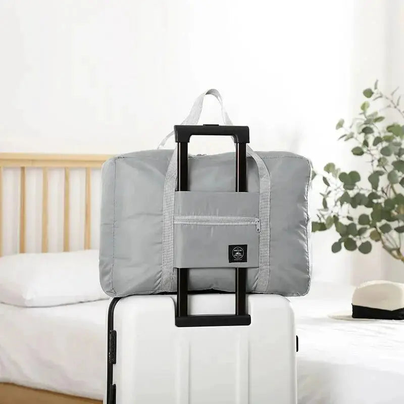 Simples Market gray foldable travel duffel bag with front zip pocket on a white carry-on suitcase.