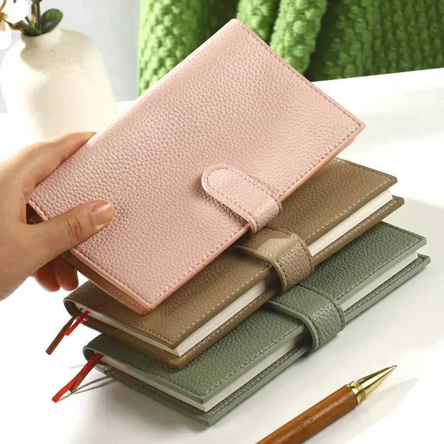 A6 weekly planner notebook with pink leather cover and strap, stacked with four planners in beige, brown, and green.