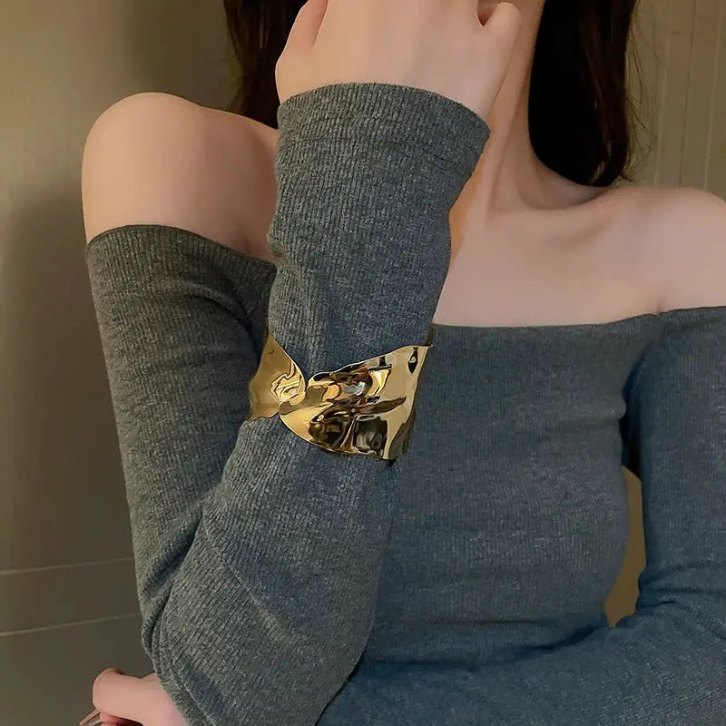 Gold-tone metal cuff bracelet with geometric twist on a woman's wrist, worn over a gray off-shoulder top.