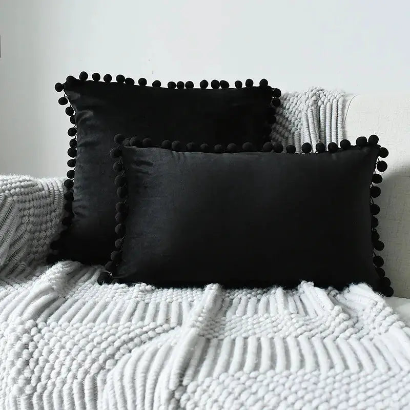 Two velvet cushion covers with black pom-pom fringe on a light sofa.
