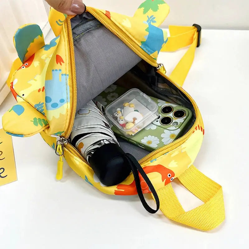 Cute yellow kids backpack with cartoon dinosaurs; open main compartment showing a phone and a small snack container