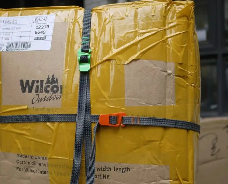 Luggage strap with quick-release buckles securing a yellow taped cardboard package; orange and green buckles on dark gray webbing.
