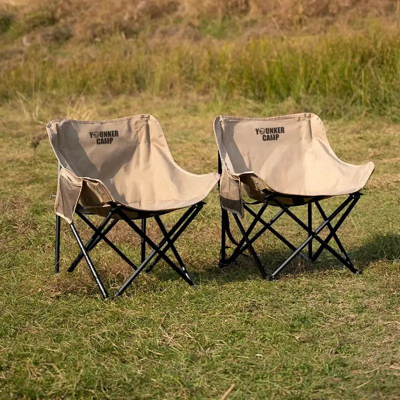 Two beige folding camping chairs with black metal frames and YOUNKER CAMP branding, outdoor grass setting
