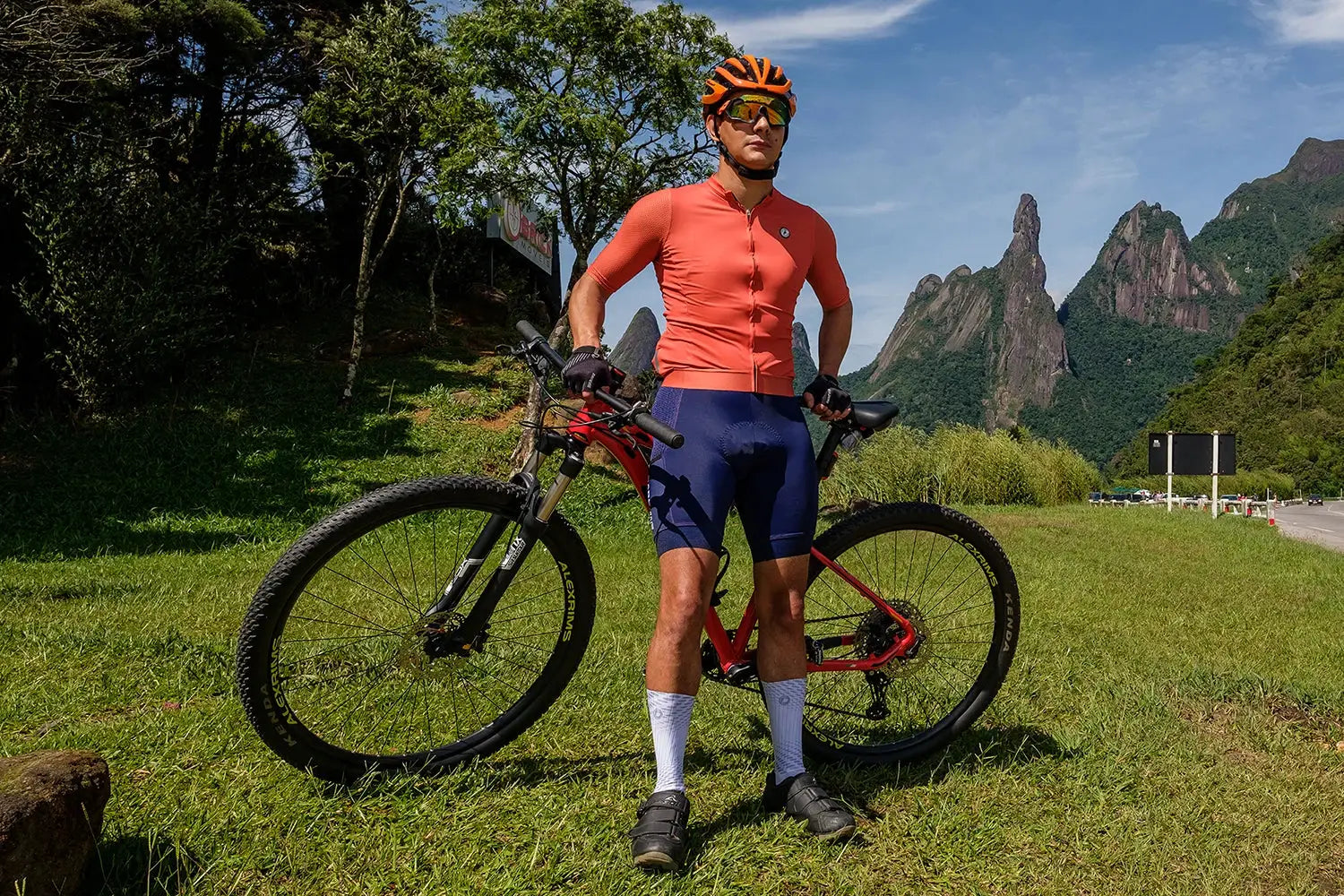 Cycling jersey by Simples Market, orange DAREVIE top with shorts, helmeted rider beside red mountain bike in field.