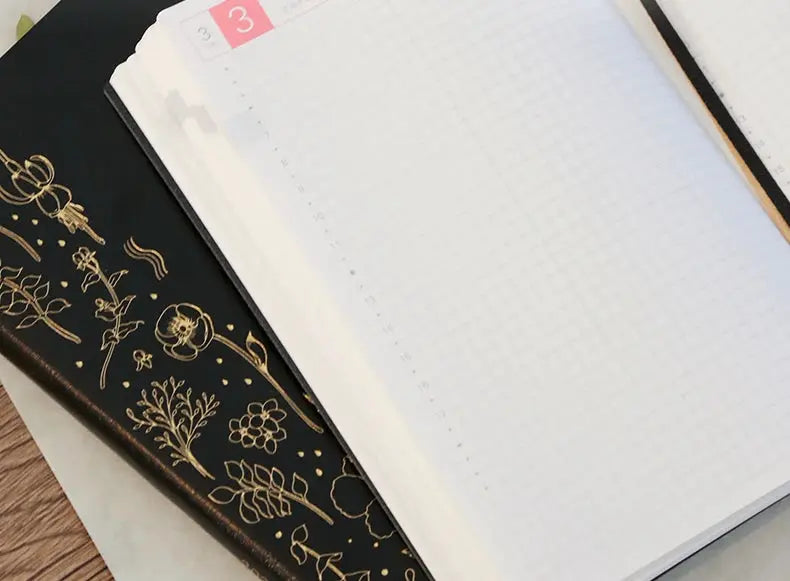 Kinbor notebook planner with black faux-leather cover and gold floral design beside an open white grid notebook.