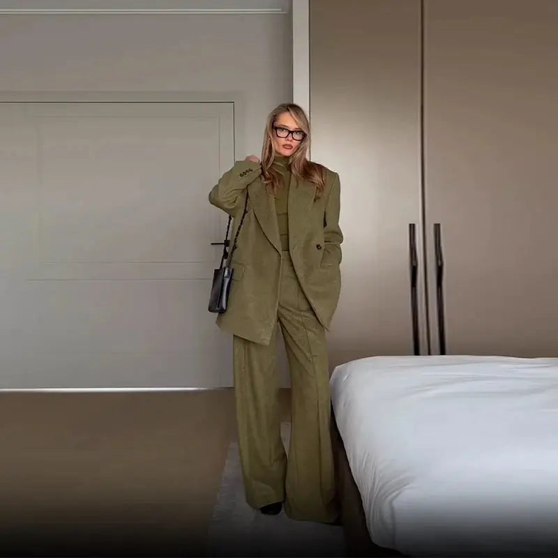 Olive green women's blazer and matching straight-leg pants suit with shoulder pads, styled with a black bag indoors