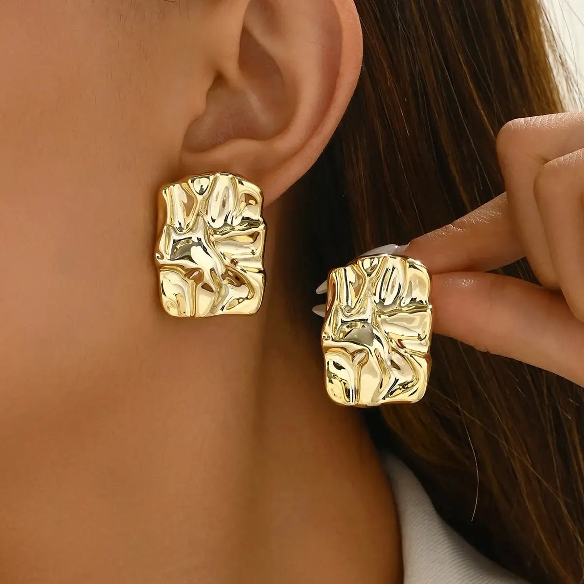 Two gold-tone geometric irregular stud earrings shown on ear and in hand