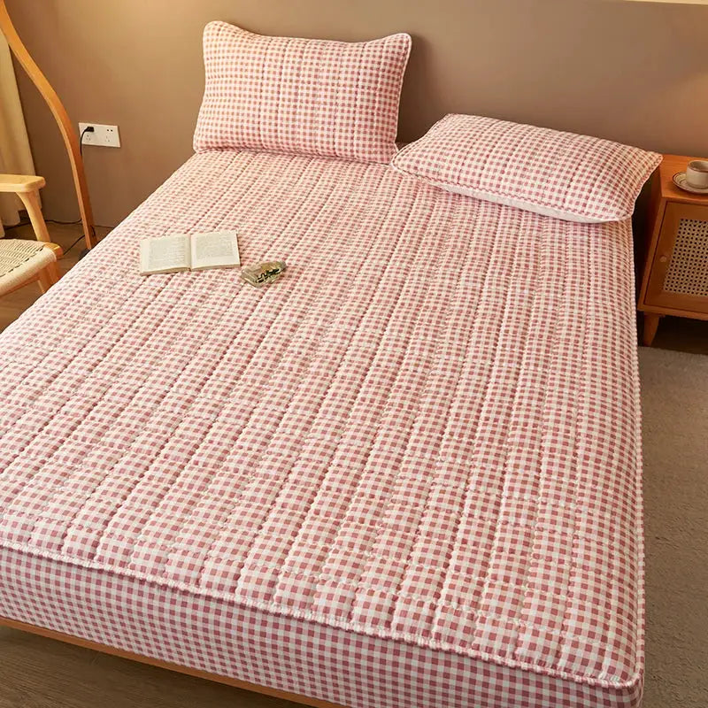 Quilted pink plaid mattress protector with two matching pillows by Simples Market on a bed