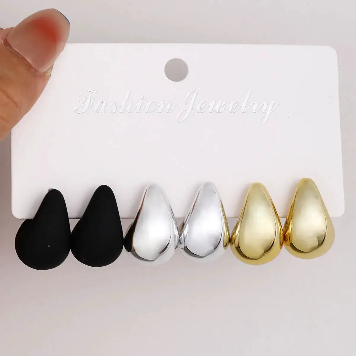 Six teardrop stud earrings in black, silver, and gold on a white fashion jewelry card