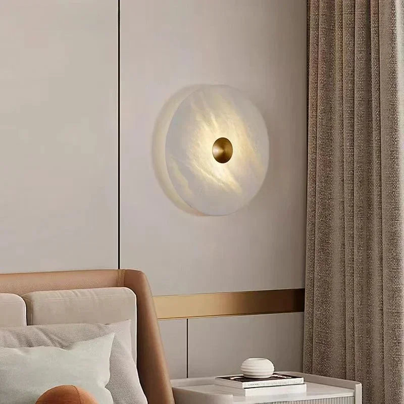 Wall lamp with natural white marble disc and brass center, mounted above a bed in a modern bedroom.