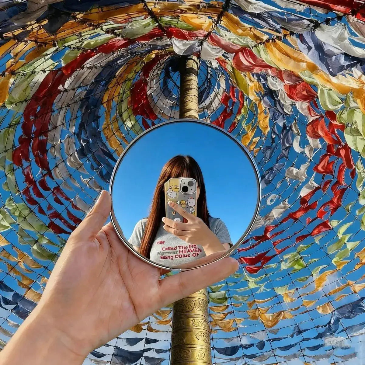 Travel handheld selfie mirror by Simples Market; round convex mirror reflecting person taking a photo against blue sky.