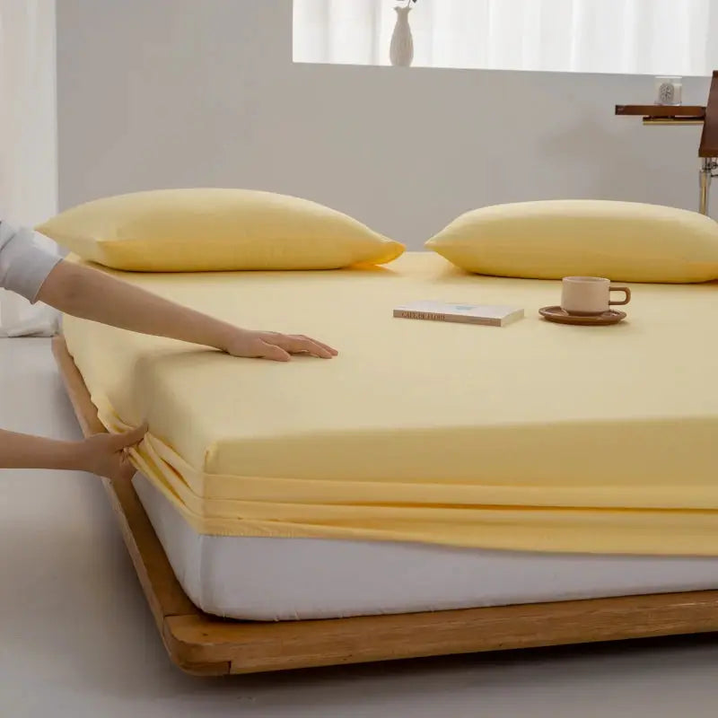 Soft cotton fitted sheet by Simples Market in pale yellow with elastic edges on a bed with two pillows, a book and a cup