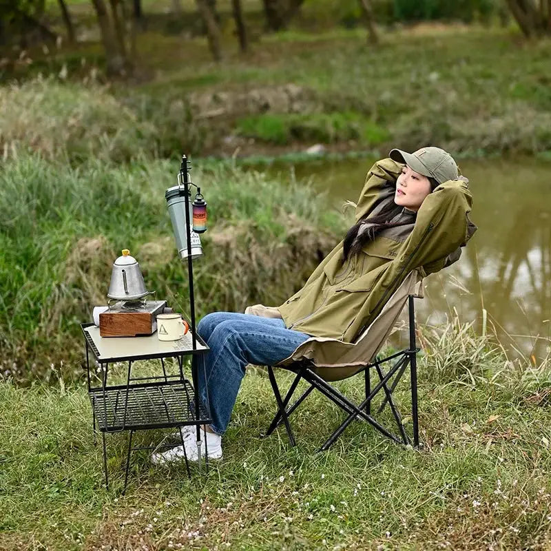 Outdoor folding moon chair in olive green with a black metal frame beside a camping table and kettle.