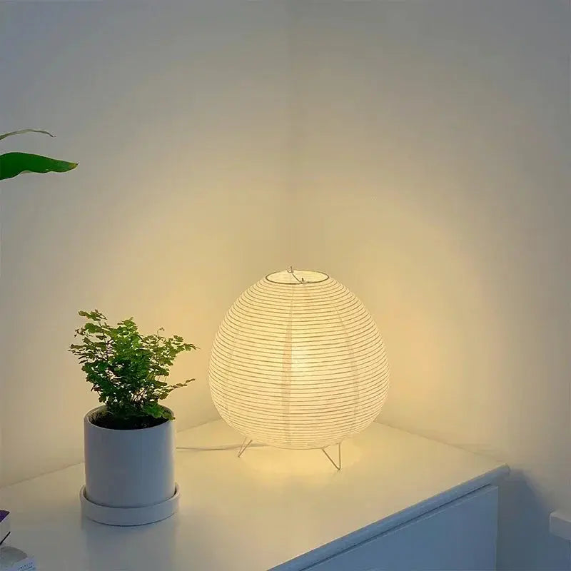 Japanese lantern LED table lamp with white rice paper shade glowing softly on a white surface beside a potted plant