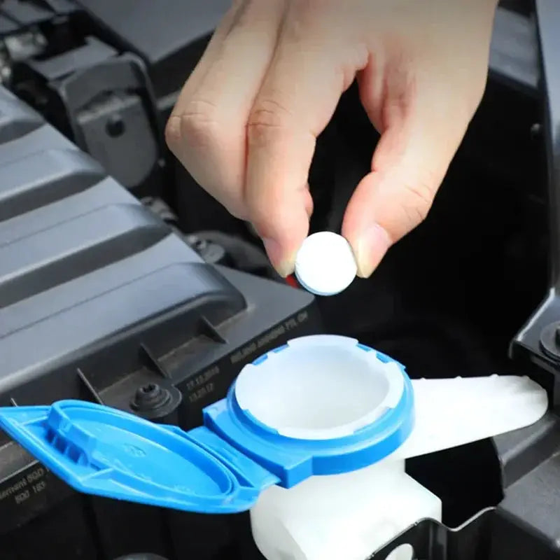 Car windshield washer tablet being dropped into blue washer fluid reservoir; white round tablet