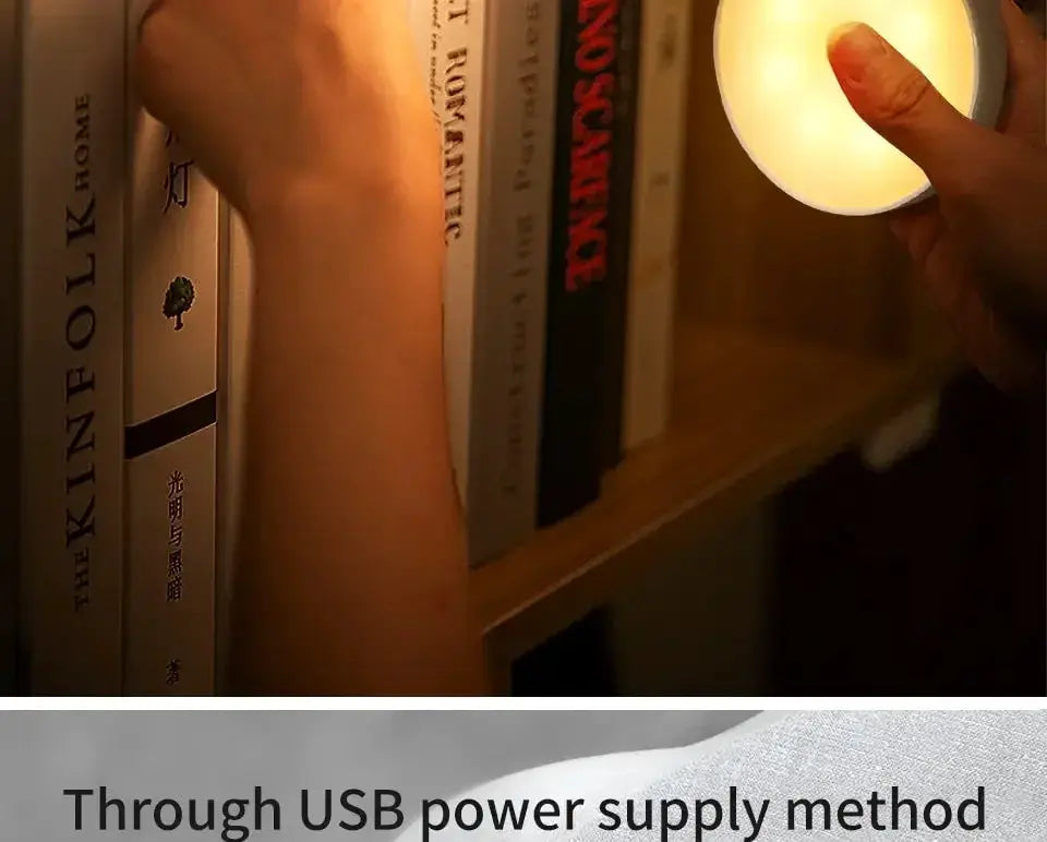 USB-rechargeable LED motion sensor night light; round puck lamp glowing warm white beside books on a shelf.