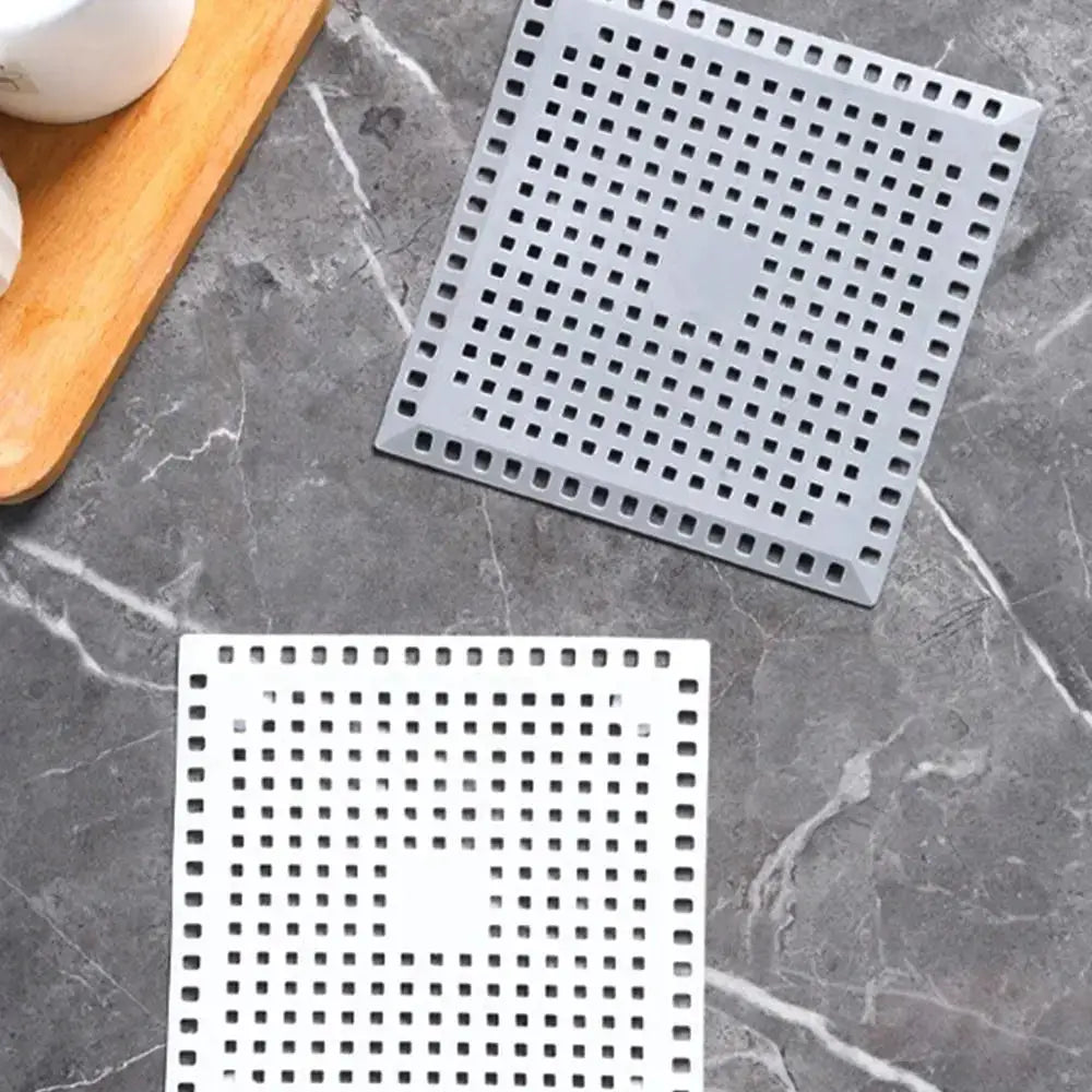 Two square kitchen sink drain strainers with perforated grids in silver and gray on a dark countertop.