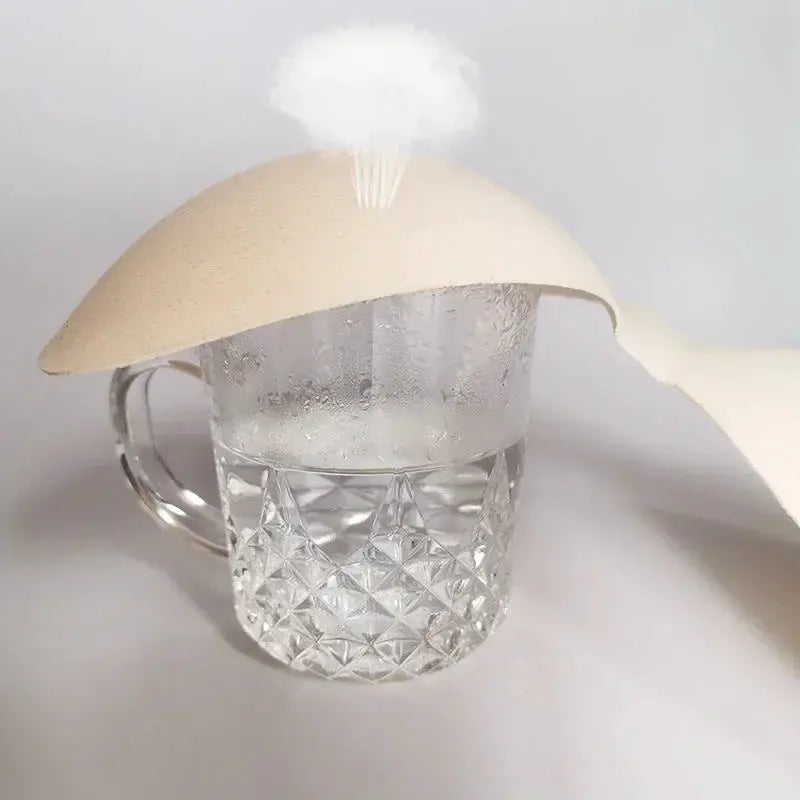 Beige sponge foam bra pads for swimsuits, one pair, displayed on a glass mug