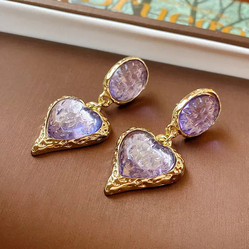Two gold-tone heart-shaped drop earrings with purple resin insets and textured frames
