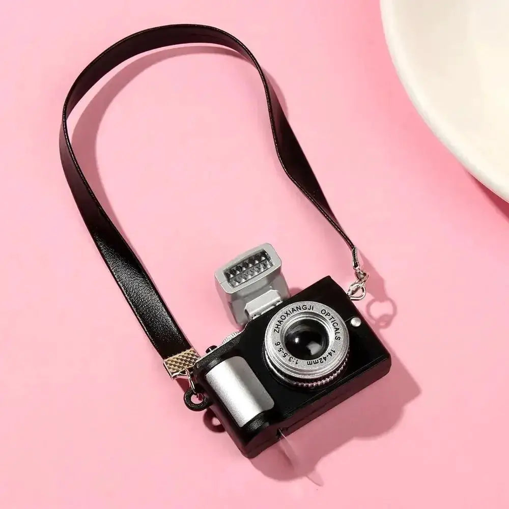 Dollhouse miniature digital SLR camera by Simples Market with black body, silver lens, and attached strap